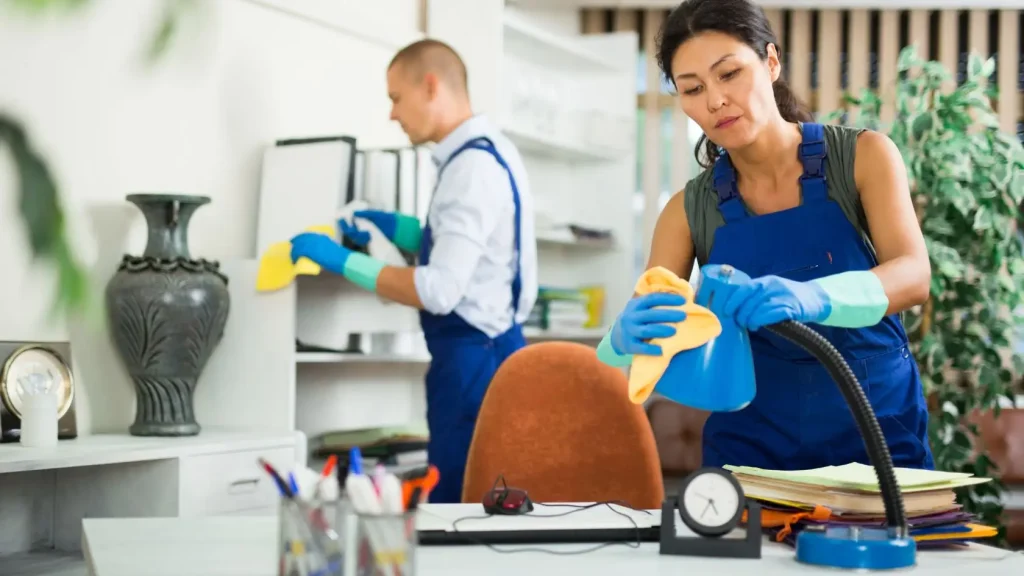 Two professional cleaners in blue overalls and protective gloves cleaning a bright, modern office space.