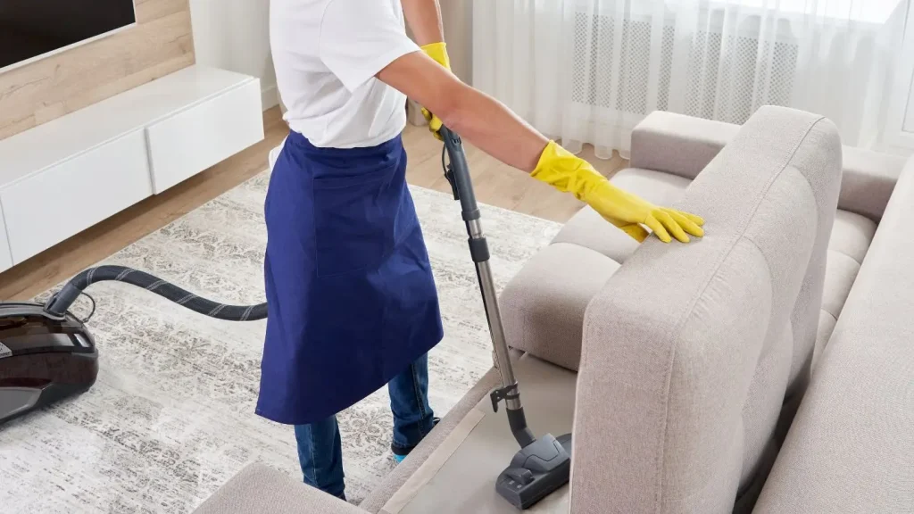 A professional cleaner wearing a blue apron and yellow rubber gloves vacuuming a fabric couch in a living room.